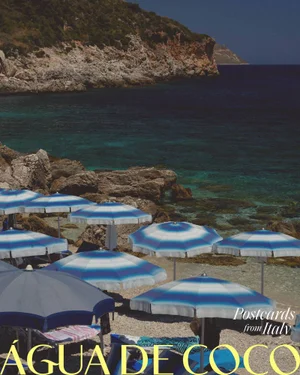 Blue and white striped beach umbrellas overlooking a rocky Mediterranean cove and turquoise sea.