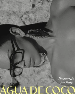 Monochrome close-up portrait of model Maria Araujo wearing square glasses and large statement earrings against textured rock.
