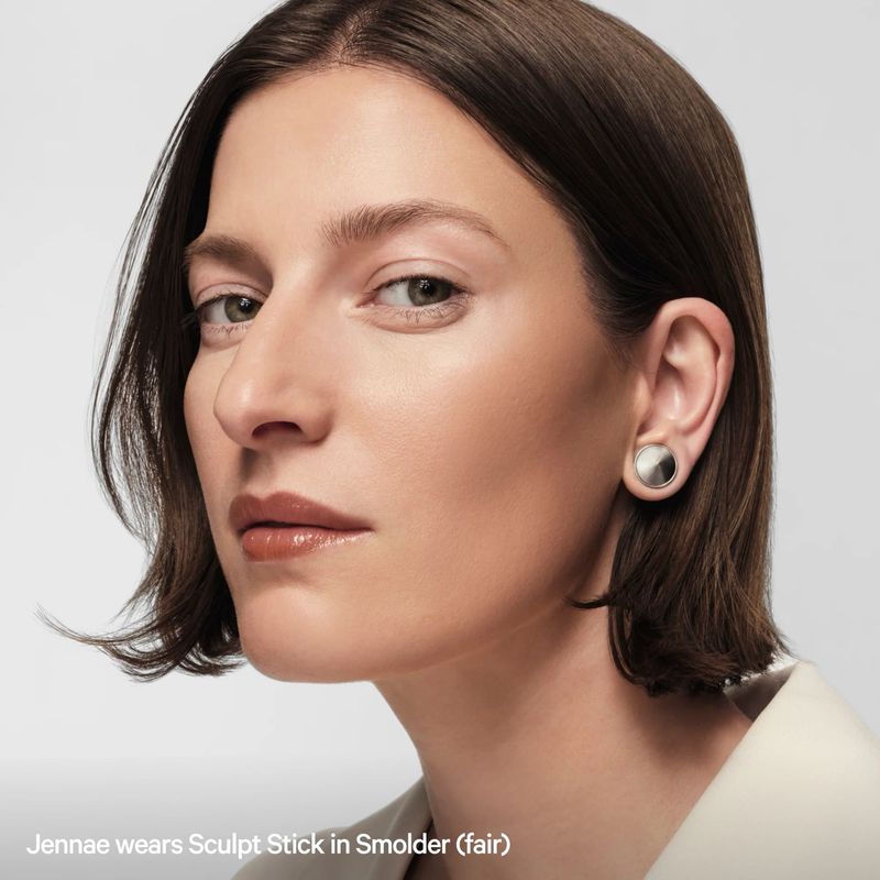 Close-up of a brunette adult female with fair skin, wearing sculpt stick on face and silver stud earrings against white background.