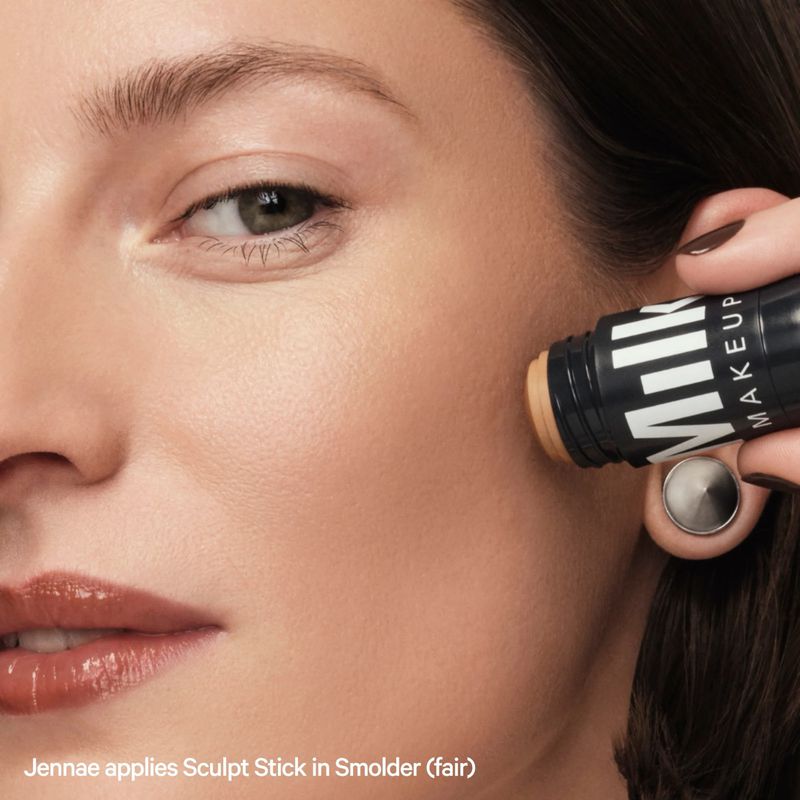 Close-up of a light-skinned woman applying face makeup with a Milk Makeup stick, showcasing beauty and cosmetic product application.
