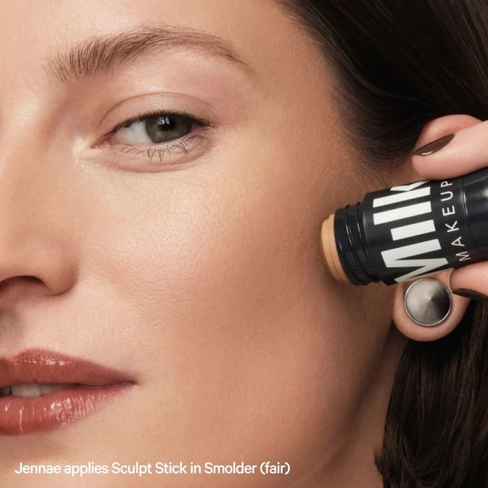 Close-up of model Jennae applying Milk Makeup Sculpt Stick cream contour to her cheek against a warm studio setting.