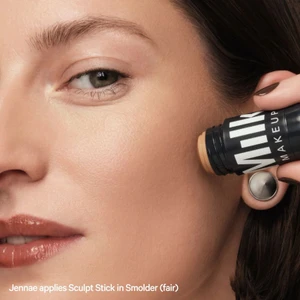 Close-up of model Jennae applying Milk Makeup Sculpt Stick cream contour to her cheek against a warm studio setting.