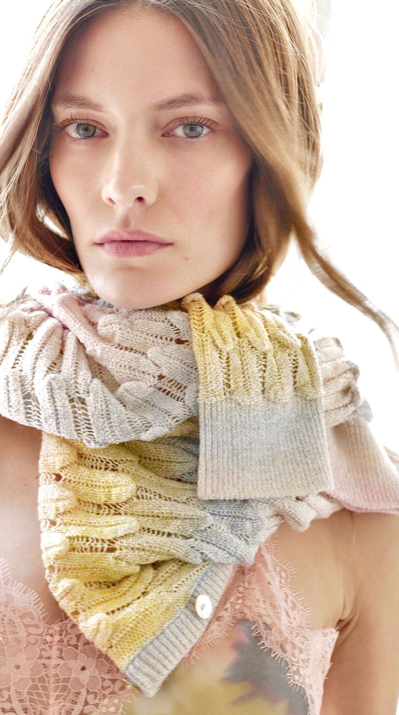 Close-up portrait of a woman with long hair, wearing a knitted scarf with pastel colors, posing in a bright, indoor environment.
