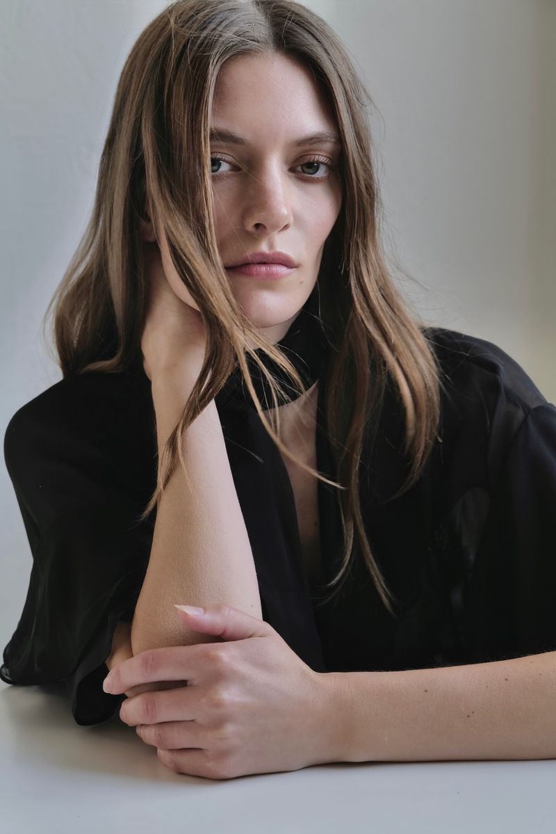 Close-up portrait of a Caucasian woman in a black top resting her head on her hand, looking directly at the camera against a plain background.