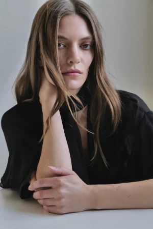 Close-up portrait of a Caucasian woman in a black top resting her head on her hand, looking directly at the camera against a plain background.