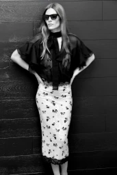 Model in black sheer blouse and patterned skirt posing against a dark wood plank wall for LeSuperbe.