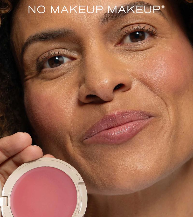 Close-up of a woman's face with light makeup, holding a container of blush. Beauty product advertising, skin details are clearly visible.