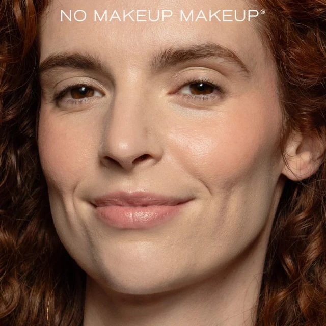 Close-up portrait of a woman with natural makeup and curly red hair smiling at the camera in an indoor studio setting.