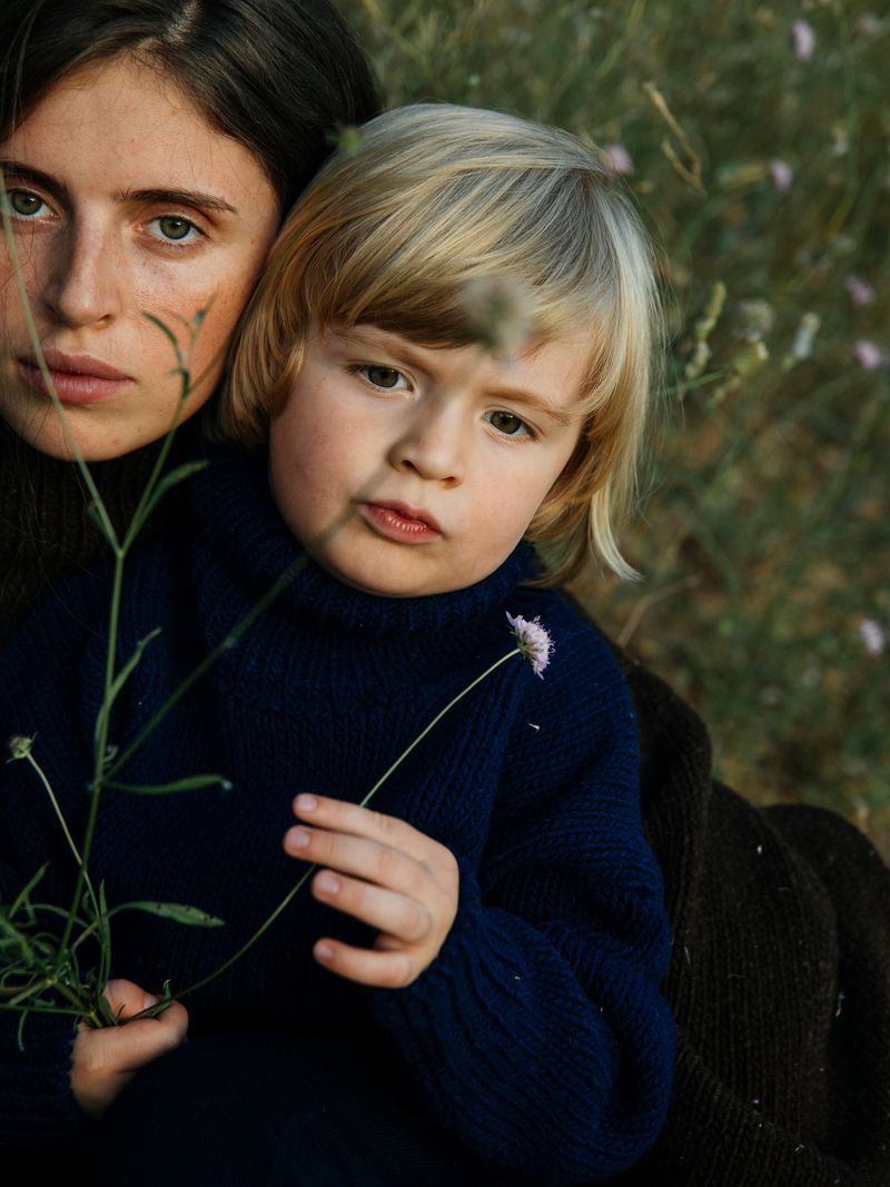 Portrait of an adult woman and a child, both with fair skin and dark clothing, holding sprigs of flowers in a grassy outdoor setting.