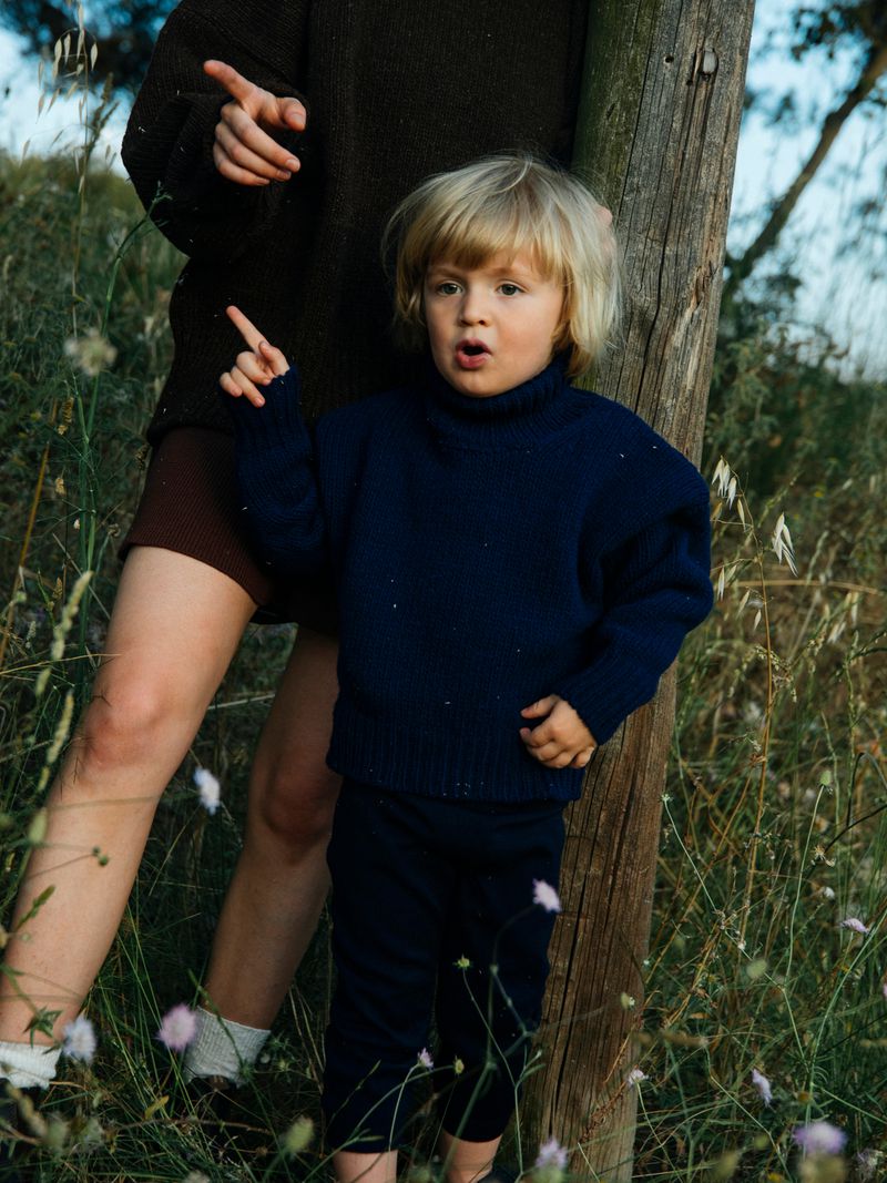 A young child with blond hair stands outside in a blue sweater, next to a woman's legs, both near tall grass and a wooden post on a sunny day.