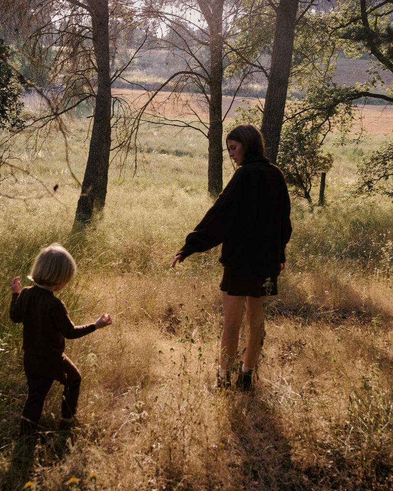 Young child and woman in dark clothing in a sunlit grassy field surrounded by trees.