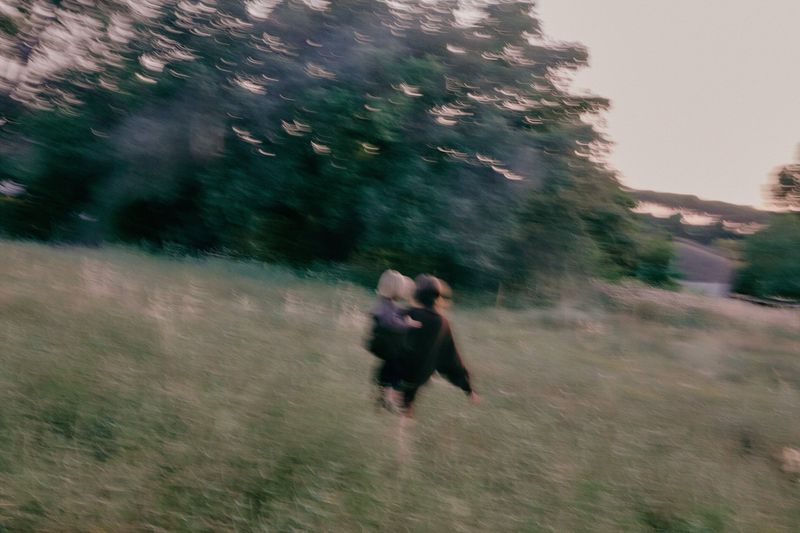 Motion-blurred photograph of a person carrying a child while moving across a grassy meadow bordered by dark trees.