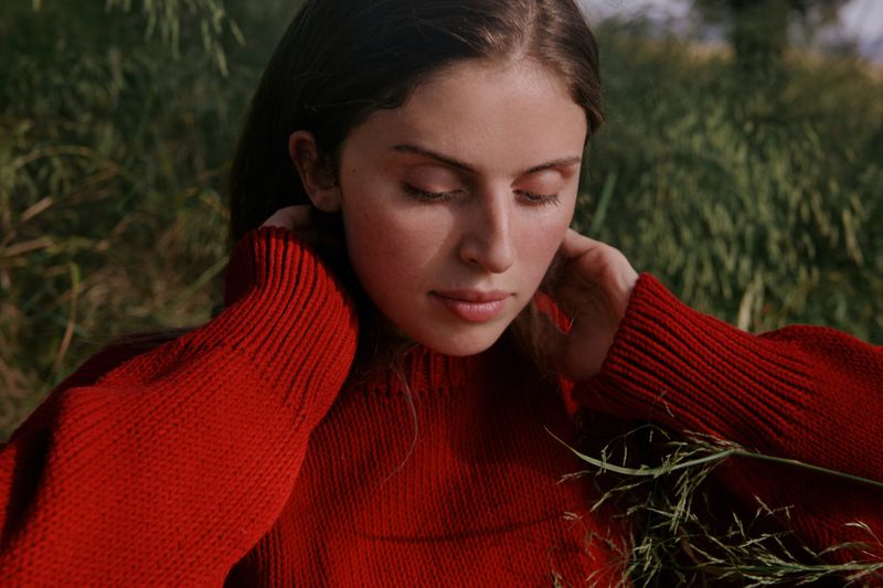 Close-up of a young woman in a red sweater posing outdoors in natural light. The model's eyes are closed, hands near her face, and surrounded by green foliage.
