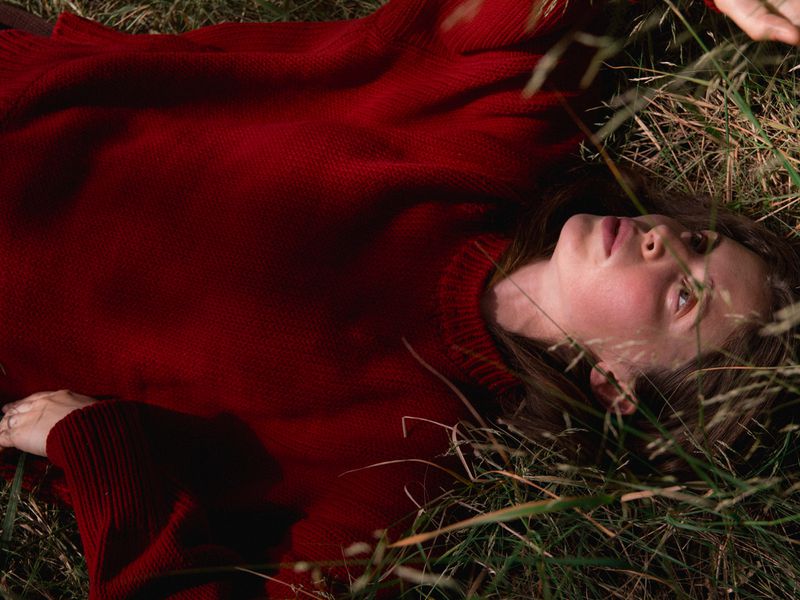 A woman lies in a field of grass, gazing upward; she is wearing a red knit sweater and the scene is illuminated by dappled natural light.