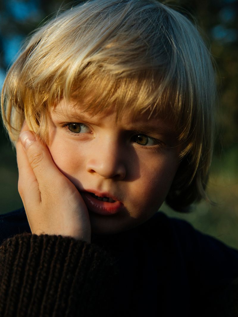 Close-up portrait of a young boy with blond hair touching his face with a concerned expression, set against a blurred outdoor backdrop.