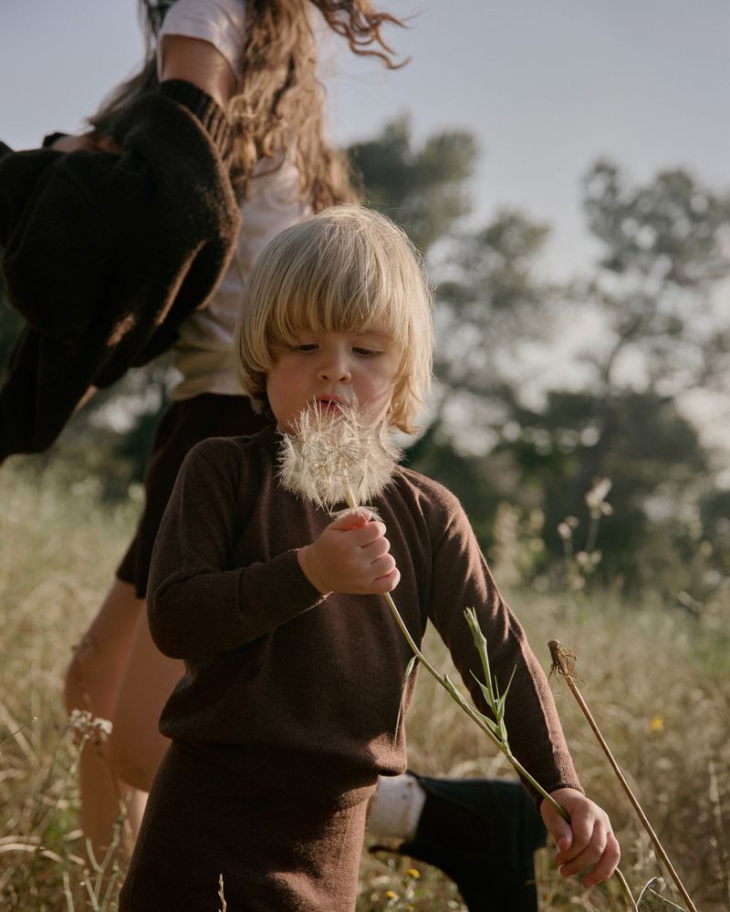 Young child with blonde hair holds dandelion outdoors in sunny meadow. Mother stands in background. Brown matching outfits.