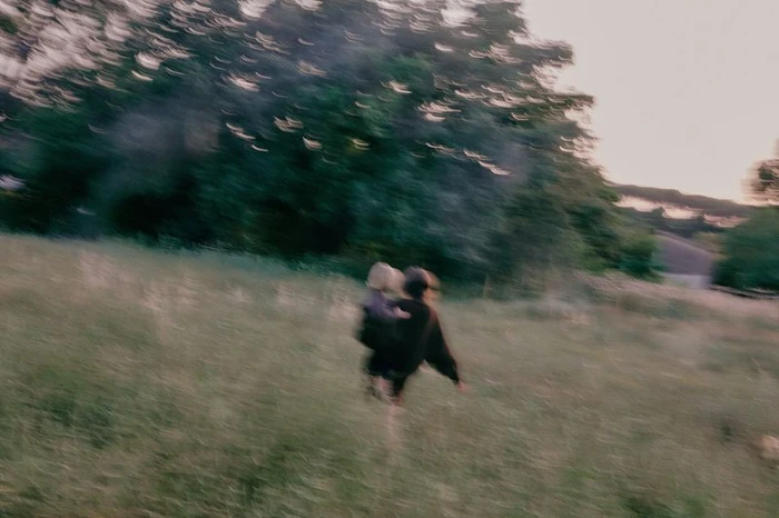Motion-blurred photograph of a person carrying a child while moving across a grassy meadow bordered by dark trees.