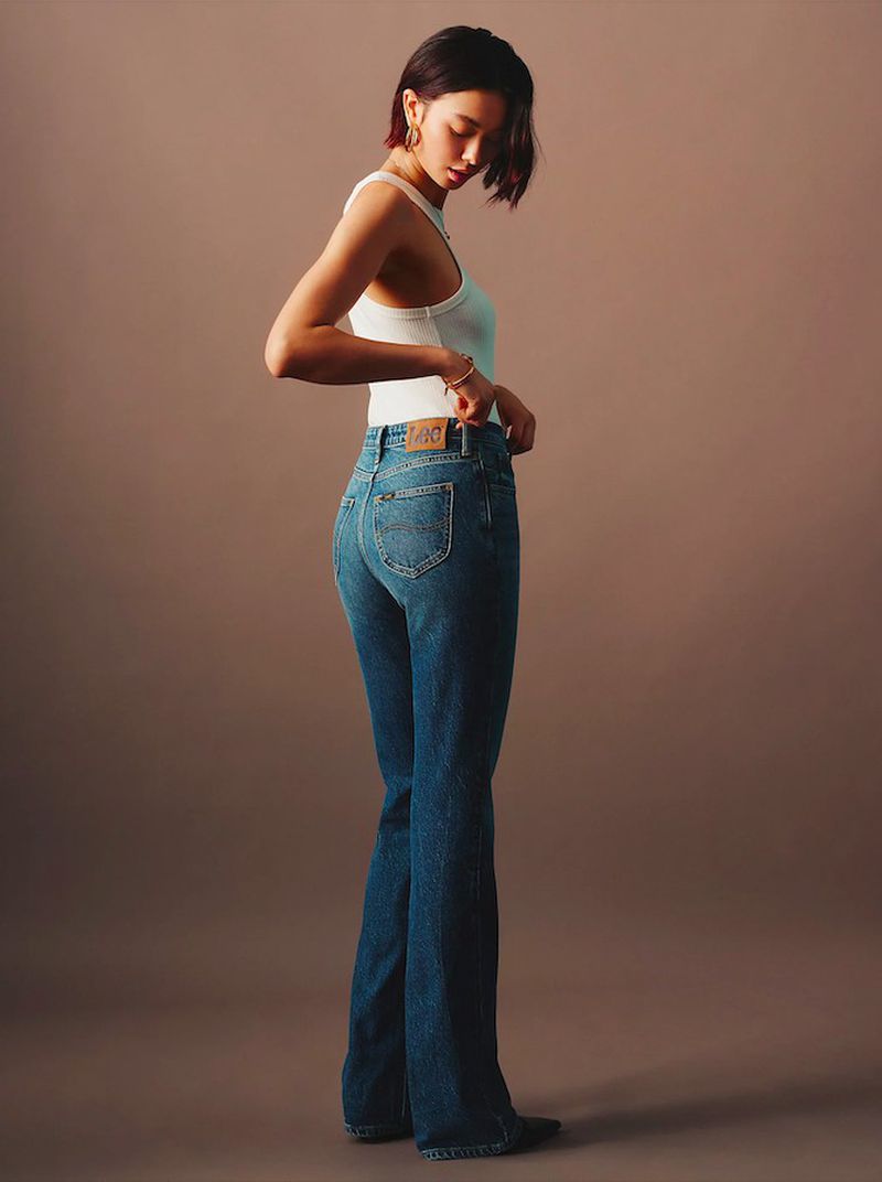 Brunette woman in a white tank top and blue jeans posing against a neutral background in a fashion studio shot.