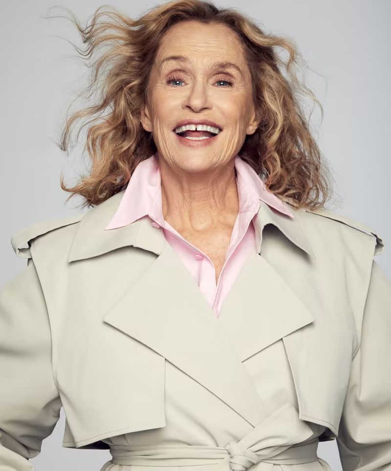 Close-up studio portrait of a smiling, mature woman with wavy blonde hair, wearing a pink blouse and light beige trench coat.