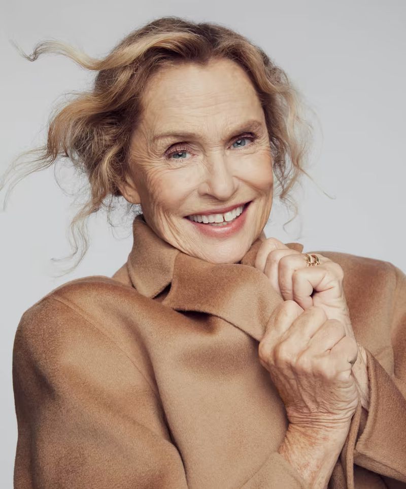 Portrait of Lauren Hutton smiling and wearing a camel coat against a seamless grey backdrop for David Jones.