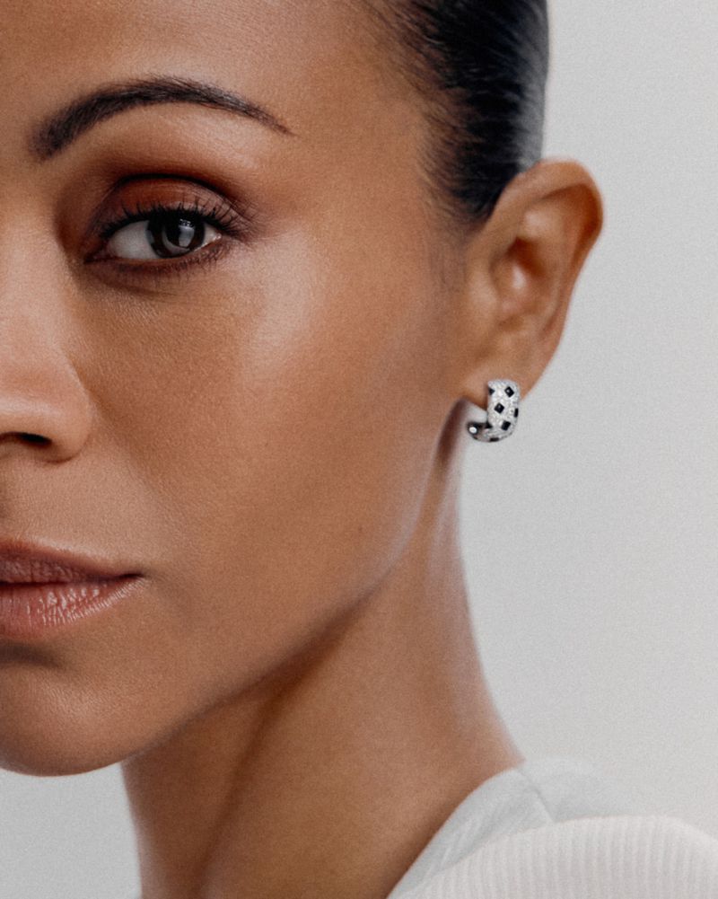 Close-up shot of an adult female with light brown skin wearing a silver hoop earring with black spots, in a neutral indoor setting.