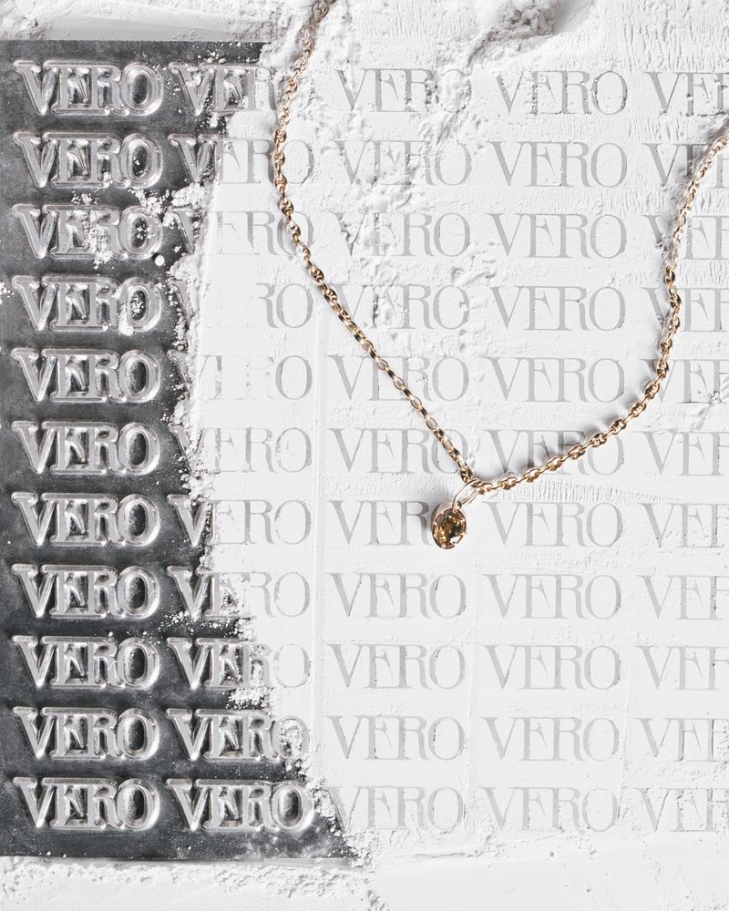 Elegant gold necklace with a solitaire pendant displayed on a textured white surface with 'VERO' text imprints for a luxury product shot.