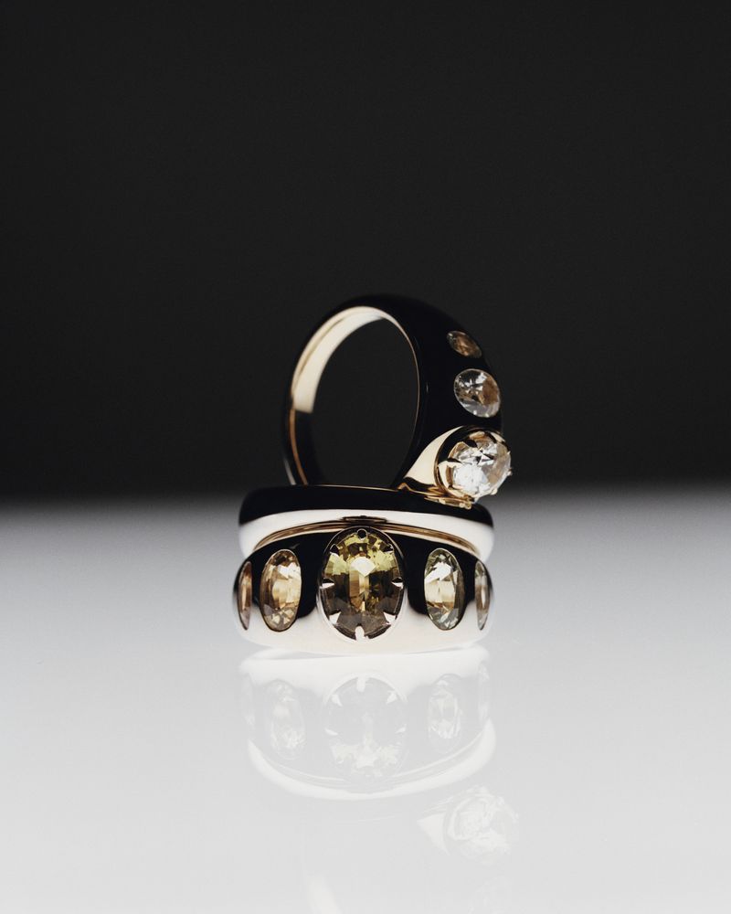 Two eye-catching gold rings are stacked together, featuring sparkling gemstones, set against a dark background with a reflective surface.