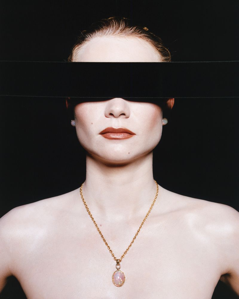Portrait of a fair-skinned woman with a black bar covering her eyes, wearing a gold necklace with a light pink pendant against a black backdrop.