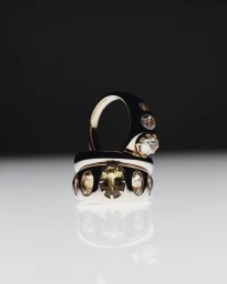 Two chunky metallic rings set with oval gemstones resting on a reflective white surface against a dark background.