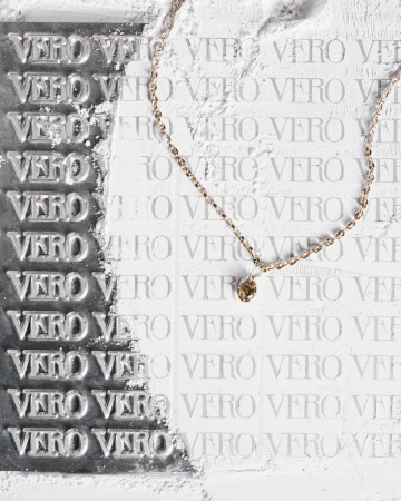 A delicate gold chain necklace with a small brown crystal pendant laid across a white background stamped with repeating VERO text.