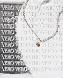 A delicate gold chain necklace with a small brown crystal pendant laid across a white background stamped with repeating VERO text.