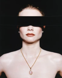 Close-up portrait of a woman with a black band covering her eyes, wearing a gold pendant necklace against a black background.