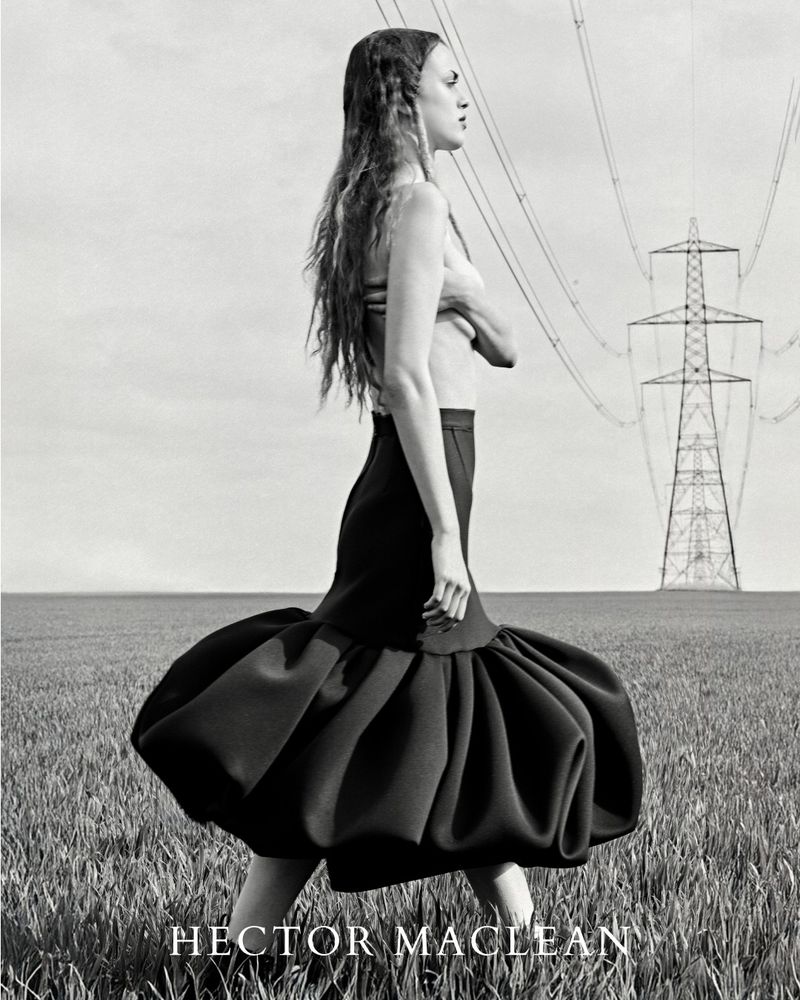 A side view of a woman walking in a field wearing a black skirt, with overhead power lines and an electrical tower in the background.