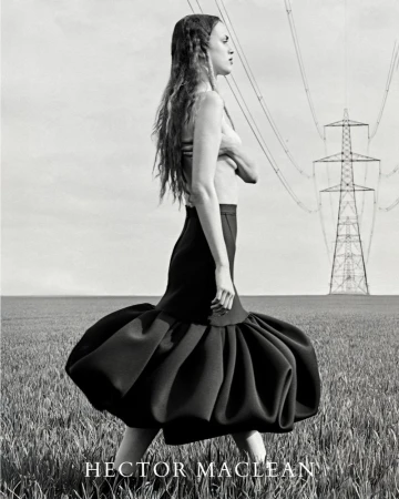 A side view of a woman walking in a field wearing a black skirt, with overhead power lines and an electrical tower in the background.