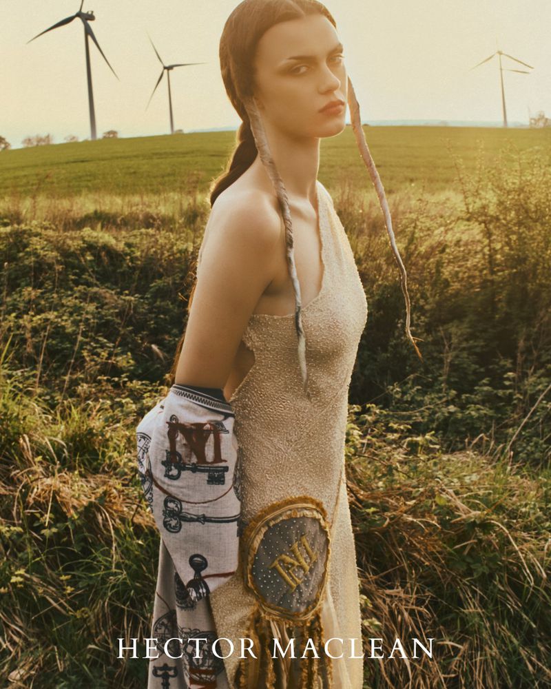 A woman in a dress stands in a grassy field with wind turbines in the background. The light is warm, and the dress has unique embellishments.