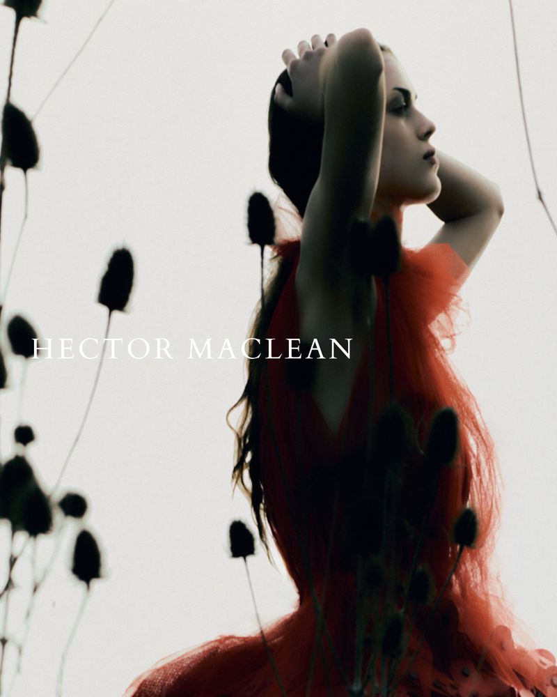 A fashionable woman in a red tulle dress poses with hands behind her head against a blurred background with tall black wildflowers.