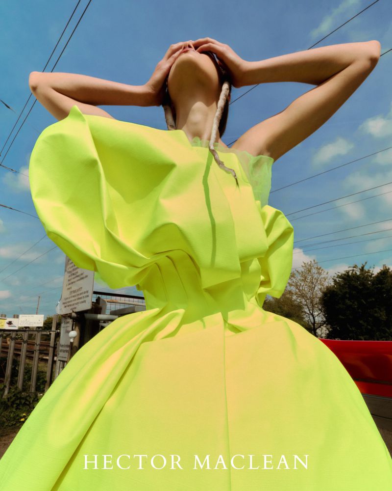 Fashion shot of a woman in a neon yellow gown, viewed from a low angle against a blue sky with powerlines and greenery.