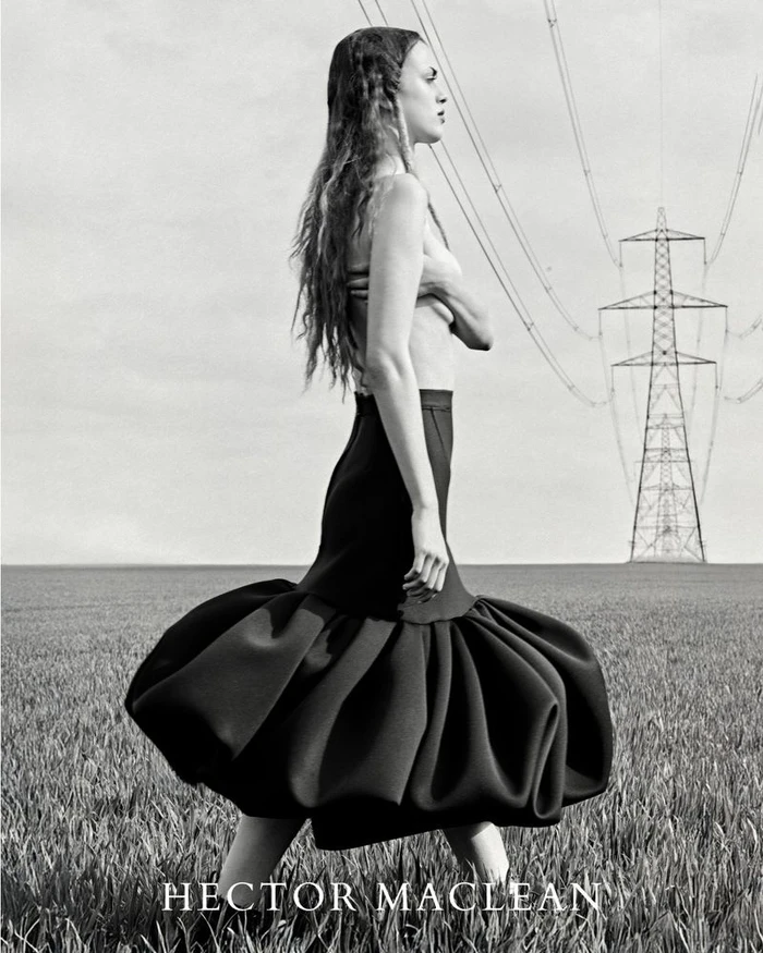 Monochrome fashion editorial featuring a model in a voluminous black skirt standing in an open field beneath utility pylons.