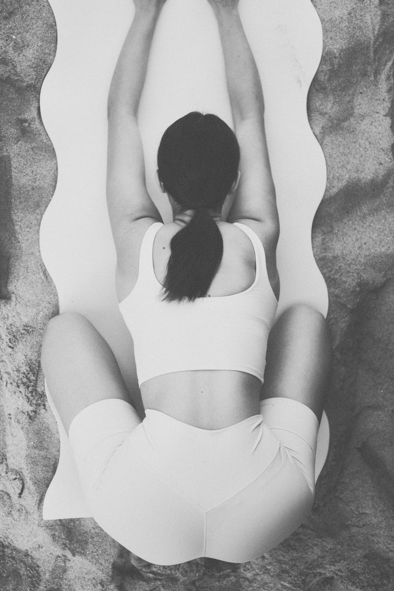 Overhead shot of a woman stretching on a white yoga mat placed on a sandy surface, wearing light-colored athletic wear.
