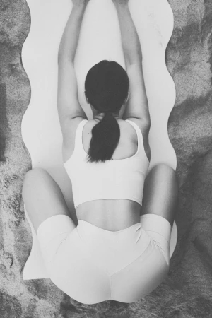 Overhead shot of a woman stretching on a white yoga mat placed on a sandy surface, wearing light-colored athletic wear.