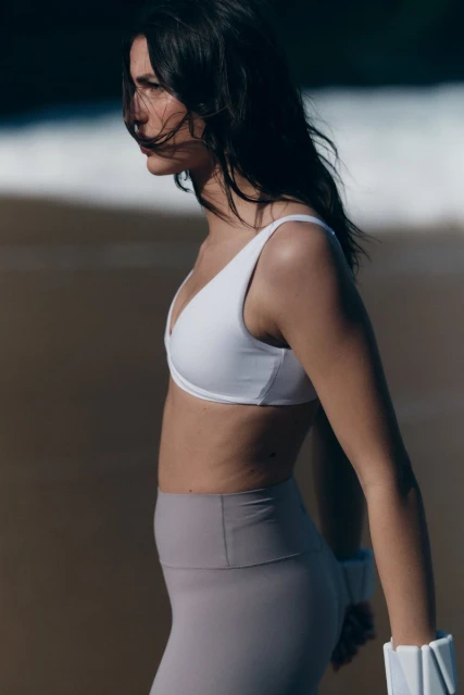A woman with dark hair stands on a beach wearing workout clothes with the ocean and sand visible in the background during the day.