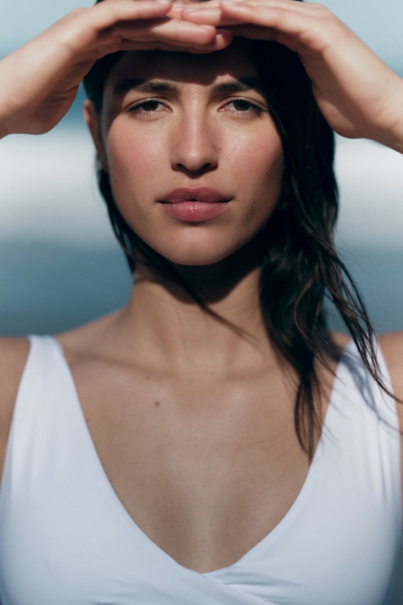 Close-up shot of a woman in a white bathing suit shielding her eyes from the sun at the beach with a blurred background.