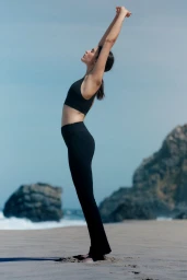 Model Adele Aldighieri wearing black activewear while stretching arms overhead on a sandy beach with rocky cliffs in the background.