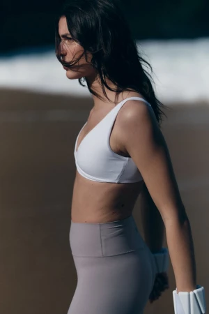 Model in a white sports bra and grey leggings standing on a sandy beach with ocean waves in the background.