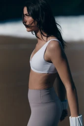 Model in a white sports bra and grey leggings standing on a sandy beach with ocean waves in the background.