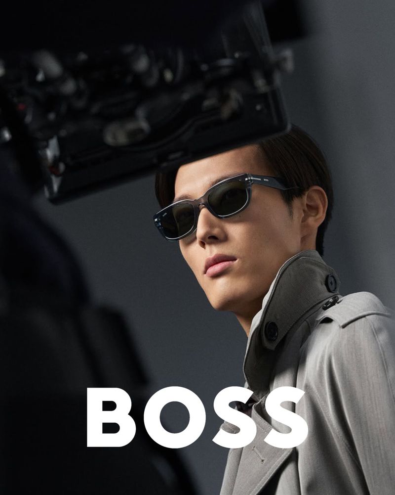Close-up of an Asian male model wearing sunglasses and a gray trench coat, posing in a photo studio with overhead lighting equipment.