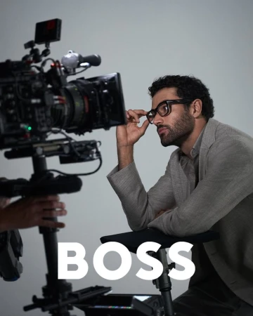 Man in eyeglasses seated near a professional video camera in a studio. He wears a grey blazer against a simple grey backdrop.