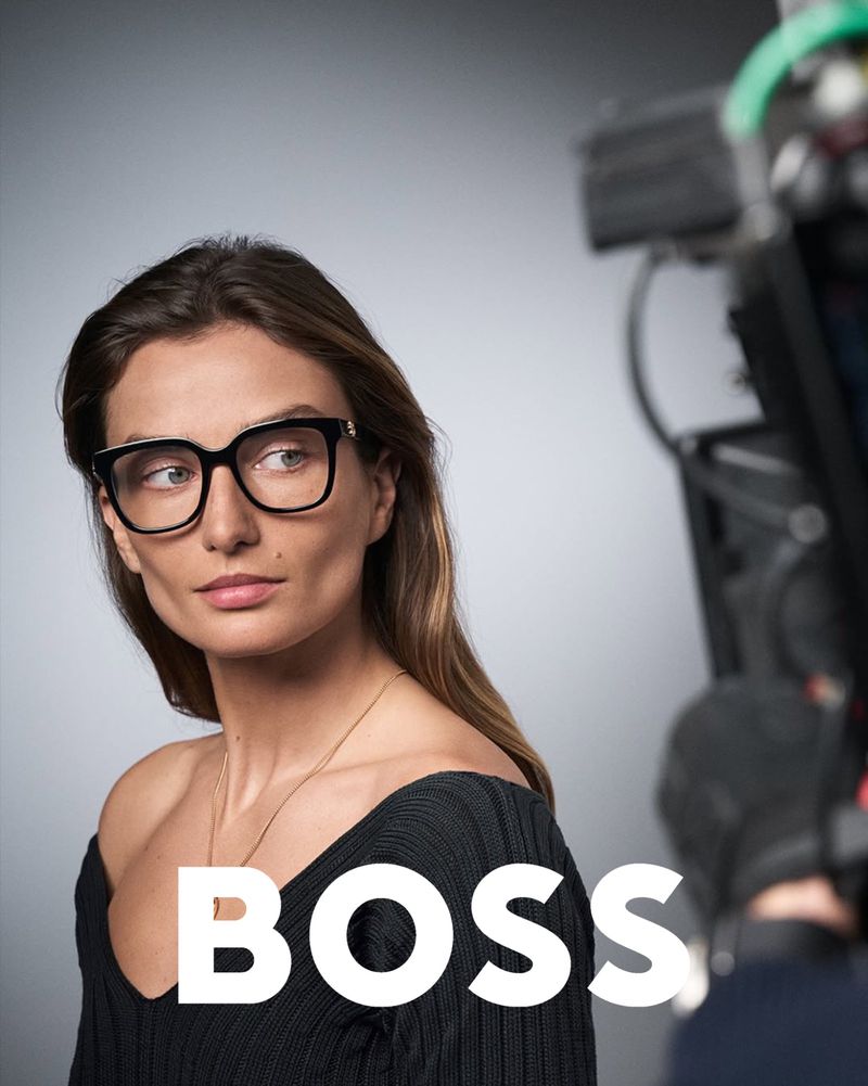 Brunette woman with glasses poses in front of a camera with the BOSS logo in the foreground, fashion or behind-the-scenes concept.
