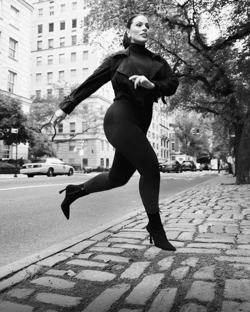 A black and white full shot of a woman running on a cobblestone sidewalk in an urban environment, wearing a dark jacket and heels.