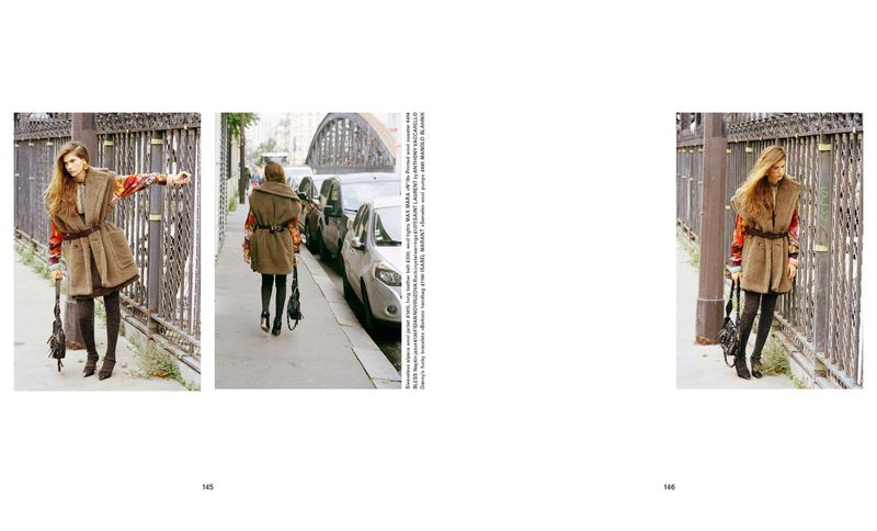 Fashion shoot features a woman in a brown coat standing on a city street next to a black iron fence in daylight, photographed outdoors.